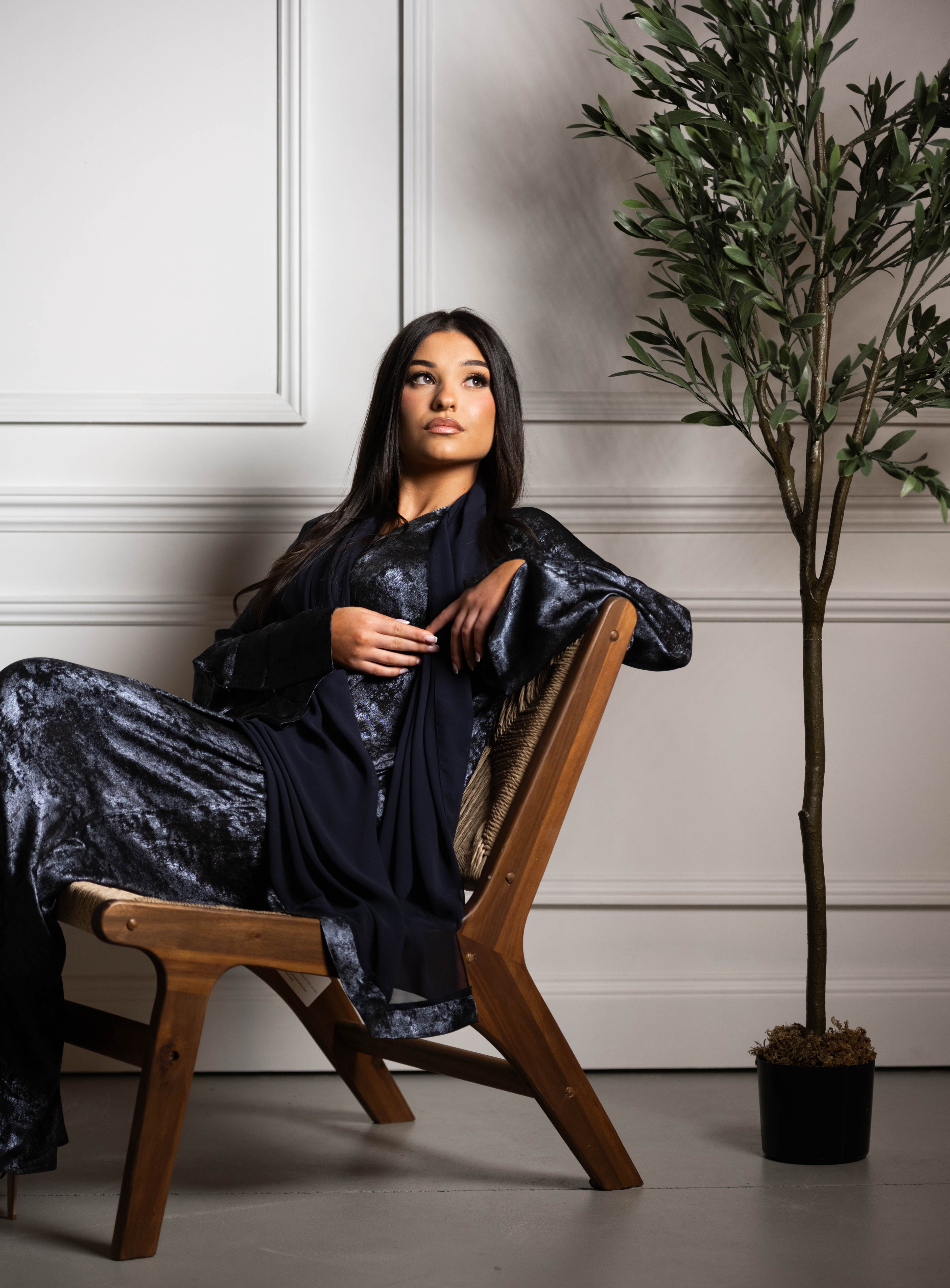 Woman in a midnight navy subtle shiny outfit sitting on a wooden chair with a plant in the background